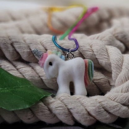 "Charming white unicorn charm with a colorful mane, nestled among intertwined beige ropes, accompanied by a hint of a vibrant, multi-colored string and a fresh green leaf.