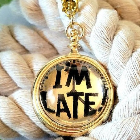 A golden "I'M LATE" pendant rests upon a tangle of creamy threads, its mirrored surface reflecting a hint of adventure, a playful accessory inspired by the whimsy of wonderland. 