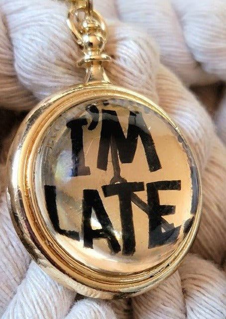  A whimsical golden pocket watch pendant boldly stating "I'M LATE" in black letters, evoking a playful nod to the classic tale of Alice in Wonderland. Its reflective surface captures the surrounding textures, adding a layer of depth and charm.