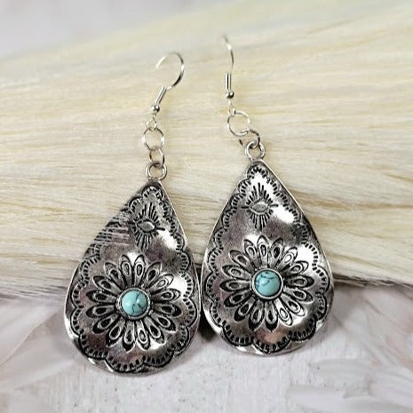 Teardrop silver earrings with floral filigree and a central turquoise stone create a striking contrast on a white furry backdrop.