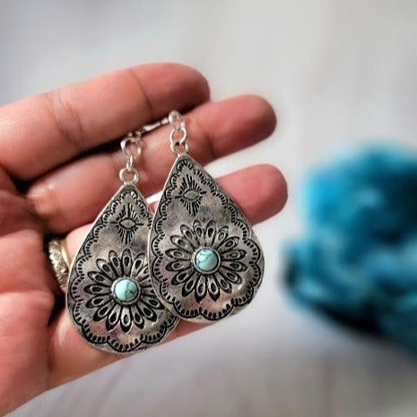 A hand presents teardrop silver filigree earrings with a central turquoise stone, showcasing intricate details against a blurred background.