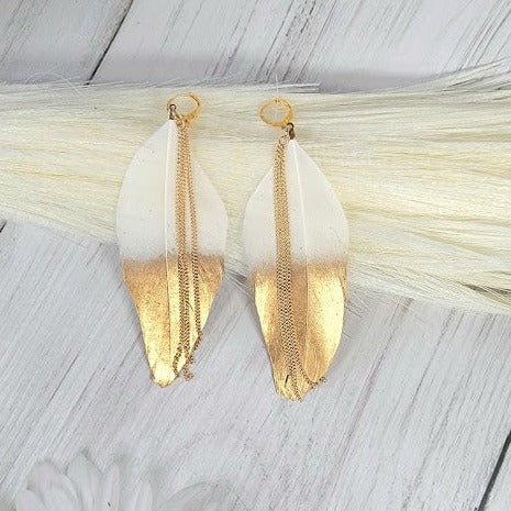 Pair of gold dipped leaf earrings with delicate chain accents, resting on a white wooden surface with a soft, ethereal background.