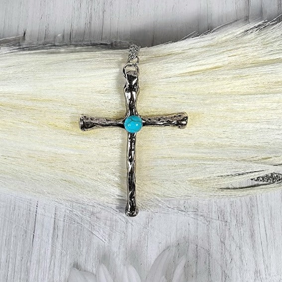 A textured silver cross pendant, adorned with a vibrant turquoise stone, symbolizes timeless faith with a modern twist, set against a plush, white backdrop.