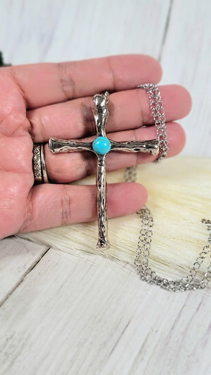 A hand cradles a textured silver cross necklace, its turquoise stone popping against the chain's shimmer, over a rustic wooden background.