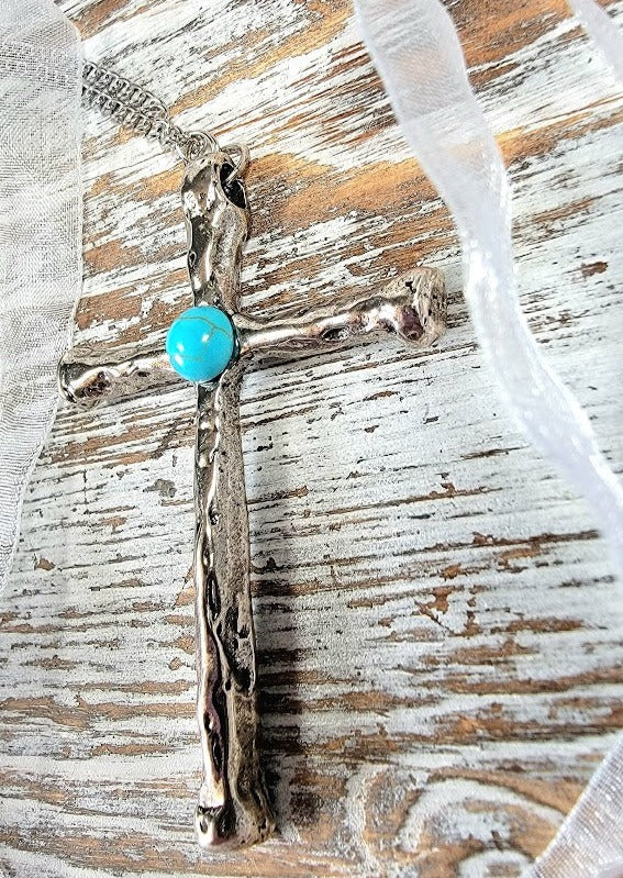 A textured silver cross pendant, centered with a bright turquoise stone, rests on a weathered wood backdrop, accented by a delicate, sheer ribbon.