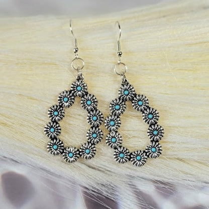 Delicate silver earrings with a geometric floral design and captivating turquoise details dangle gracefully, offering a fusion of traditional elegance and contemporary style against a soft, natural backdrop. 