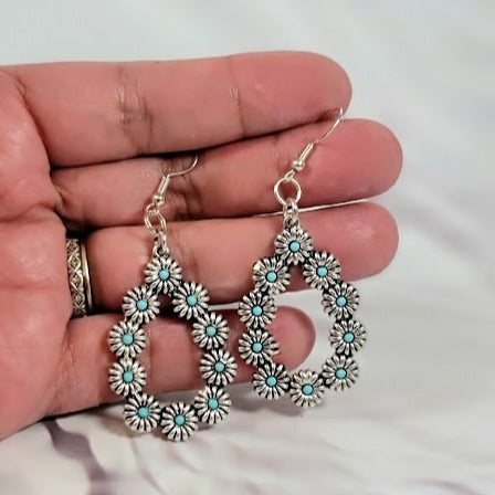  A hand presents intricate silver-toned square drop earrings with a star-like floral motif, accented by turquoise-blue dots that add a pop of color and a dash of boho elegance to the design. Ideal for a sophisticated yet playful accessory choice