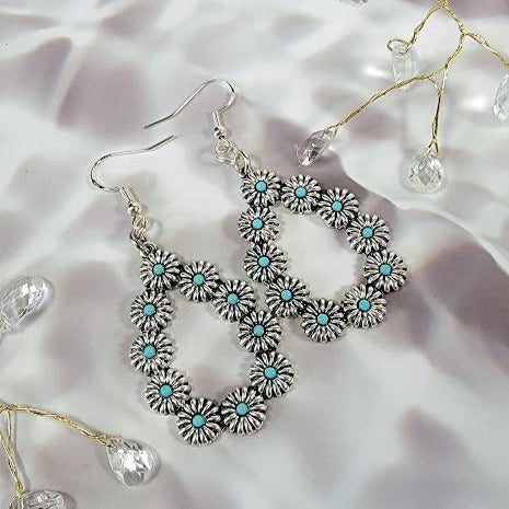 Luxurious silver earrings with an ornate, looping design, studded with turquoise-blue centers amidst petal-like details. These earrings sparkle against a satin background, offering a hint of vintage glam and bohemian allure for an elegant statement