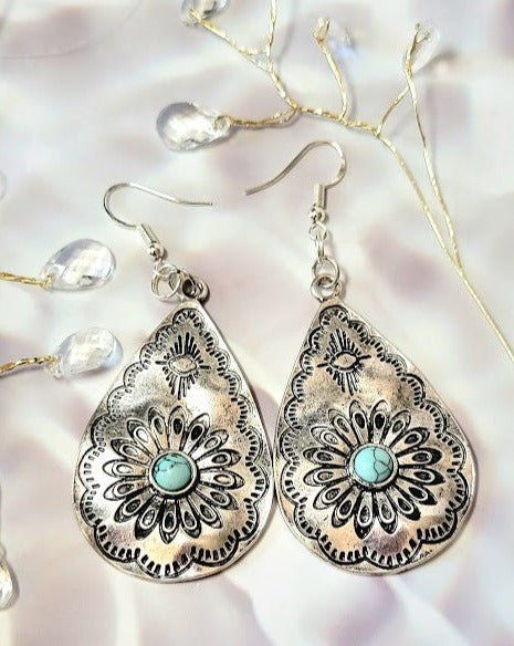 Silver filigree teardrop earrings, each with a turquoise heart, display intricate patterns and craftsmanship on a whitewashed wooden backdrop.