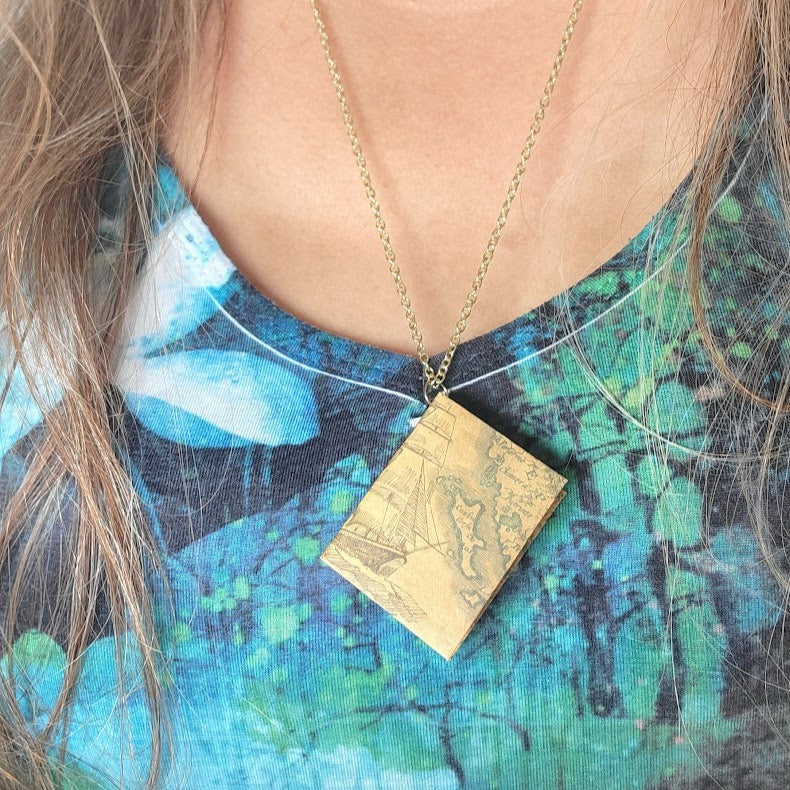 Unique square pendant with a vintage nautical map motif, set against an artistic floral dress, connected to a delicate gold chain, offers a juxtaposition of exploration and elegance.