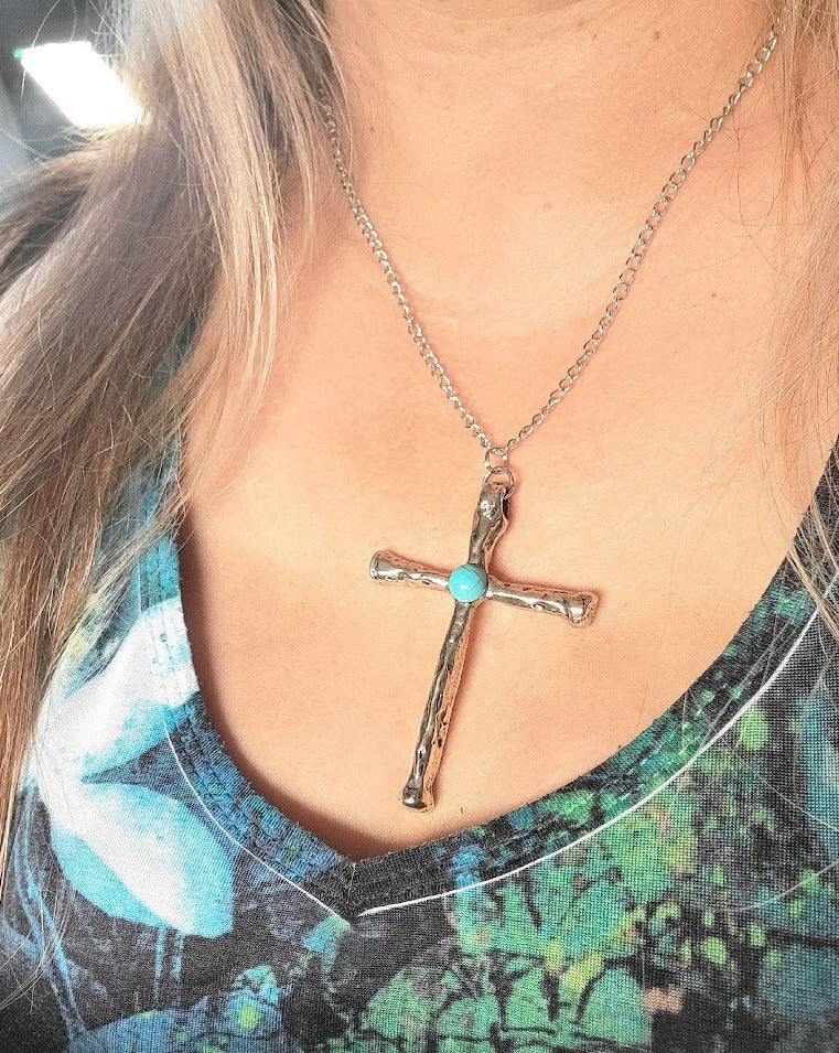 Simple yet profound, a rustic cross pendant with a turquoise center on a silver chain graces the neckline, symbolizing faith and serenity against a floral-patterned garment.