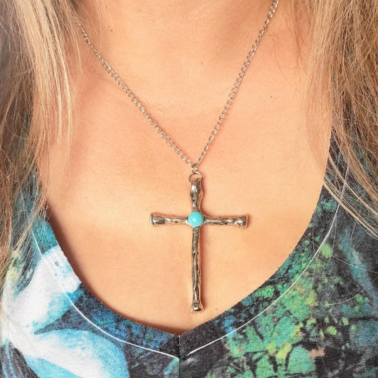 A textured silver cross with a central turquoise stone hangs on a fine chain against a floral dress, blending faith with contemporary fashion.