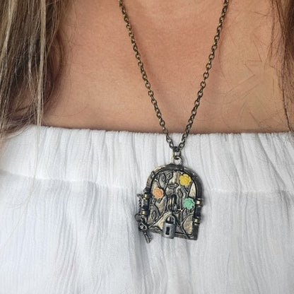 Antique-styled, ornate fairy door pendant embellished with colorful gem-like accents, hanging gracefully from a rustic chain, showcased against a delicate white blouse.