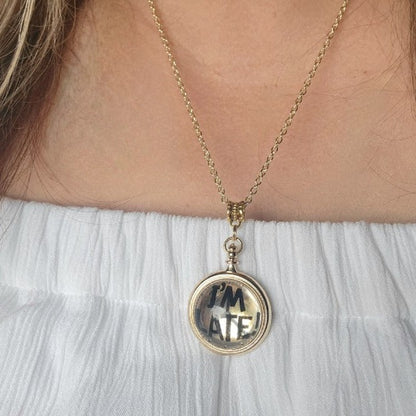 A golden "I'M LATE" pendant necklace gracefully adorns the neck of a woman, its reflective surface gleaming against her light blue attire, offering a versatile, playful accessory suitable for both whimsical and refined styles.