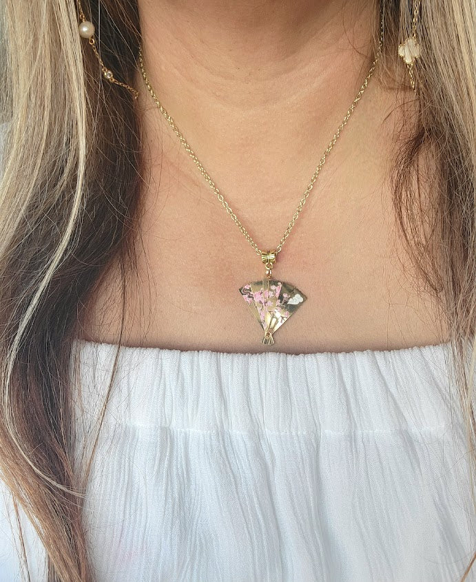 A gold fan-shaped pendant with delicate pink cherry blossoms, suspended on a fine chain, graces the neck, adding a touch of spring elegance to the white off-shoulder top.