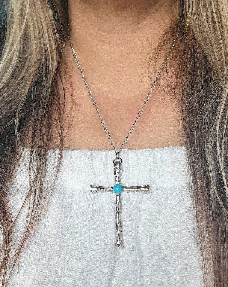 A sterling silver cross with a turquoise centerpiece is elegantly draped on a delicate silver chain, against the soft backdrop of a white dress.