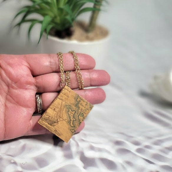 A hand displays a necklace with a vintage map pendant, channeling an adventurous spirit with old-world charm, on a delicate gold chain, amidst soft shadows.