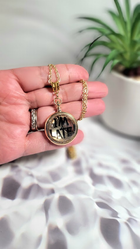 A hand delicately holds a whimsical "I'M LATE" golden pendant necklace, its playful message adding charm against a soft, shadow-patterned background, hinting at stories untold and adventures waiting.