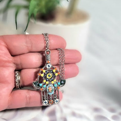 A hand presents a Hand of Fatima pendant necklace with enamel accents and a mosaic glass center against a blurred botanical backdrop.