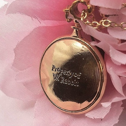 Close-up of a whimsical golden pendant with the engraving "Property of W. Rabbit" set against a blush pink floral fabric, invoking the charm of a cherished fairy tale keepsake with a personal touch. 