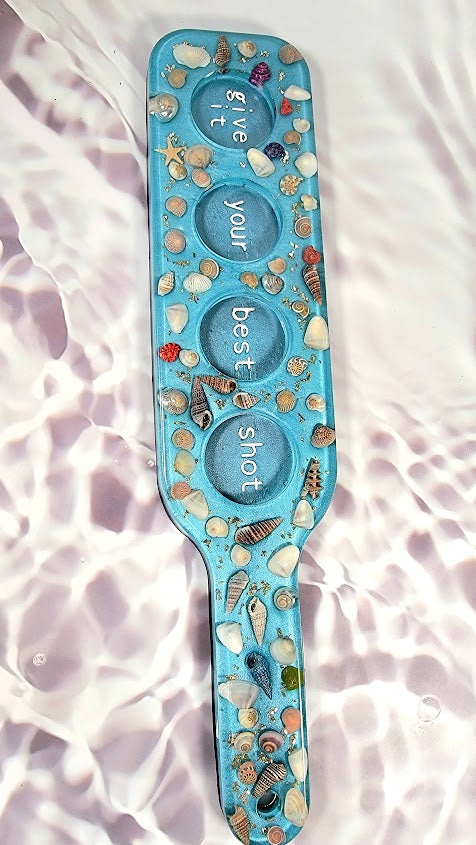 Turquoise paddle-shaped resin decor embedded with an array of seashells and sparkling embellishments, featuring the motivational words 'give it your best shot' inscribed across the surface, set against a rippled white background.