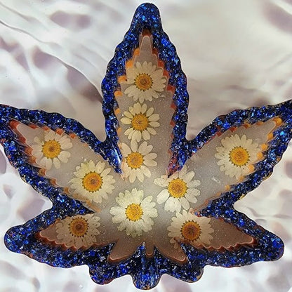 Handcrafted resin maple leaf-shaped decoration featuring a translucent core with real, delicate daisies encased within, bordered by shimmering blue glitter, presented on a textured white background.