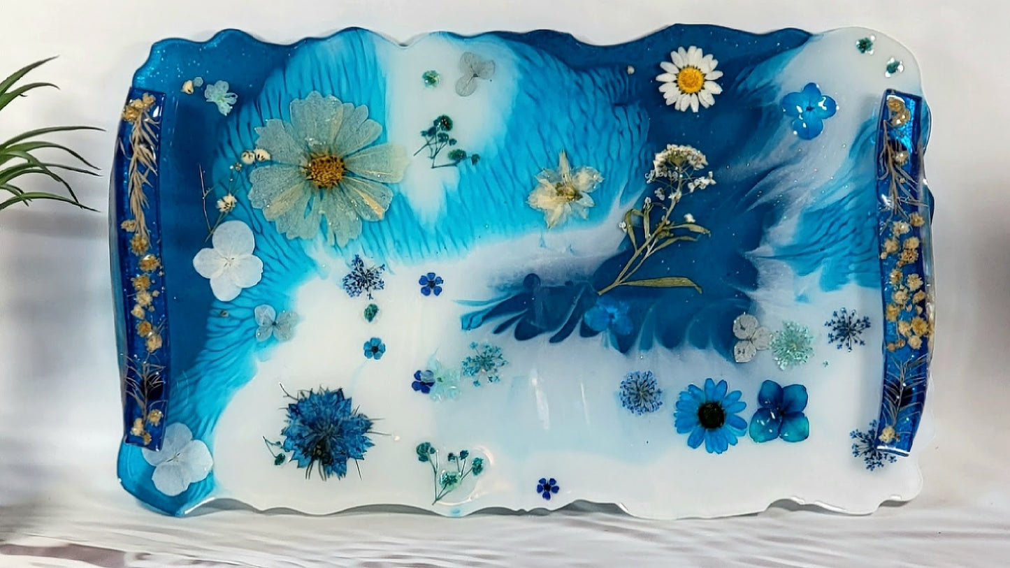 Handcrafted resin tray with intricate blue swirling patterns, adorned with an assortment of real and crafted flowers in shades of blue, white, and gold. Edges are decorated with golden accents, placed against a backdrop with tropical palm leaves.