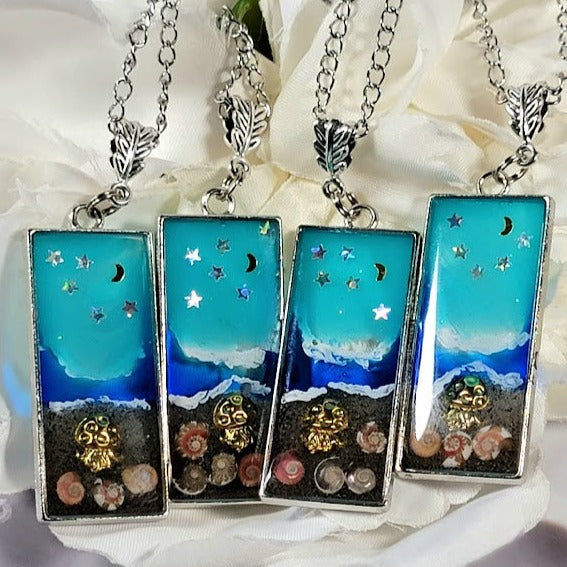 Silver necklaces featuring rectangular bezels that encase genuine seashells, set against a backdrop that resembles beach waters under a starry blue sky. All elements are sealed securely within a clear resin, presented on a soft white fabric with floral embellishments.