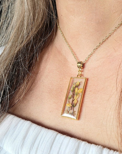 Close-up of a gold-tone necklace featuring a rectangular pendant filled with resin, showcasing preserved golden leaves and subtle embellishments, worn on a woman's neck against a white blouse backdrop.