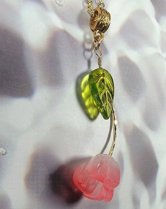 Gold pendant with a lime-green leaf charm above a translucent blush-pink rose blossom, set against a white silken backdrop.