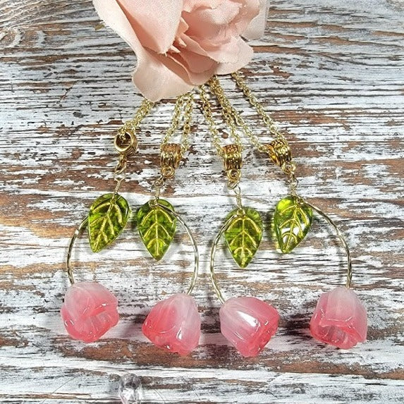 Four gold necklaces with lime-green leaf charms leading to translucent blush-pink rose blossoms, elegantly laid out beside a soft pink fabric rose on a rustic wooden surface.