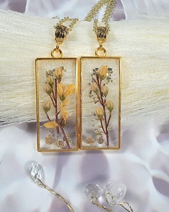 Gold-framed resin pendant earrings showcasing delicate dried flowers and shimmering gold flakes against a backdrop of white and purple silk.