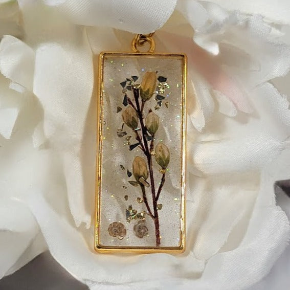 Gold-framed resin pendant necklace highlighting intricate dried flowers and subtle gold specks, elegantly juxtaposed with a soft white fabric rose.