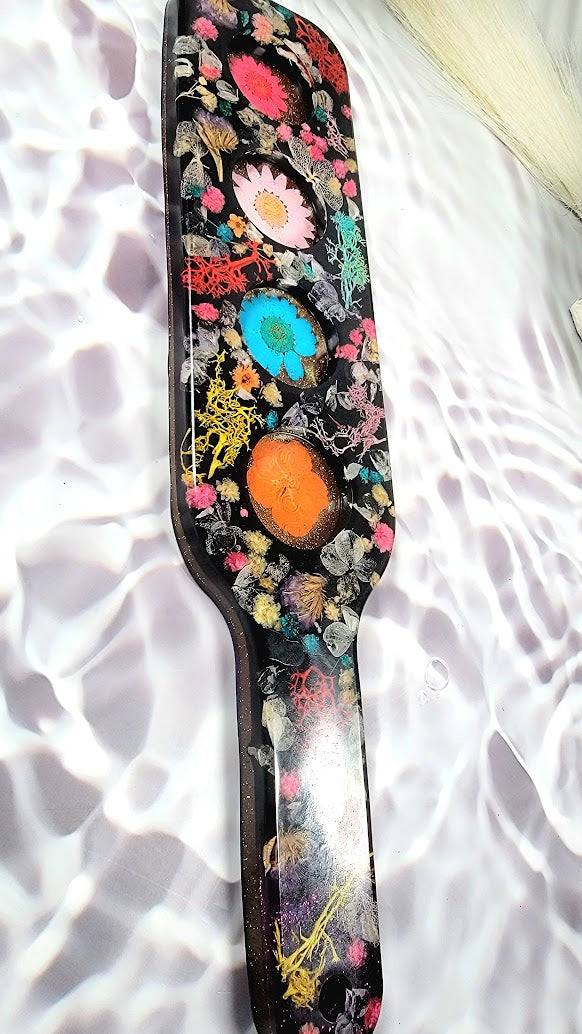 Handcrafted elongated resin art captures nature's vibrancy, a myriad of multicolored flowers, from radiant pinks to striking blues, contrasted against a deep black backdrop—the polished surface gleams under ambient light, creating a visual spectacle.