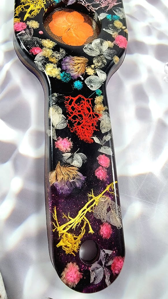 Lustrous resin piece displaying a harmonious blend of radiant flowers: cerulean blues, fiery orange, soft pink, surrounded by delicate foliage, all preserved in their natural beauty. The dark background elevates the botanical artistry, making it pop.