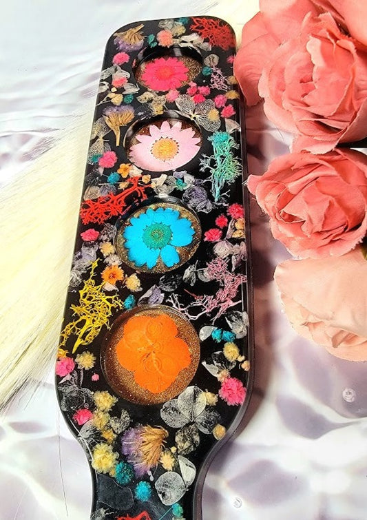 A captivating resin piece laid atop soft, white silk, intricate details of colorful flowers stand out amidst a black canvas. Soft pink roses lie adjacent, their elegance enhancing the allure. The resin's sheen captures light, offering a dynamic view