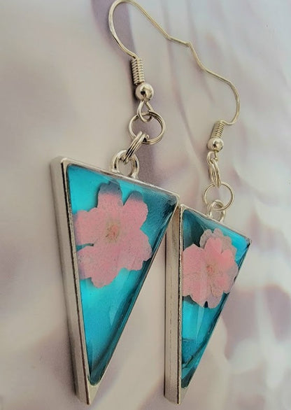 Chic, triangular resin earrings showcasing an alluring teal tint with charming pink cherry blossoms encapsulated within, paired with classic silver fishhook findings, set against a gentle, rippling white satin background.