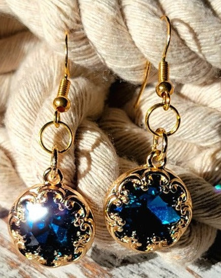 Opulent gold earrings with sparkling blue gems, framed by intricate details, exude classic elegance against a backdrop of soft, twisted rope.