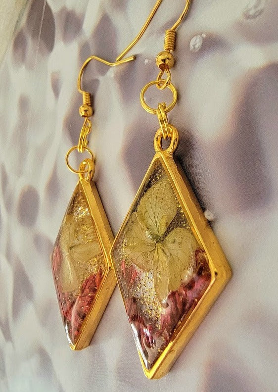 These are charming gold-framed earrings showcasing preserved flowers in amber tones suspended in clear resin, a blend of natural beauty and crafted elegance.