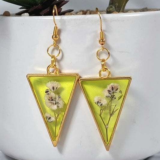 Golden triangle earrings with a vivid yellow resin and delicate white flowers inside, elegantly hung against a potted plant backdrop for a natural look.