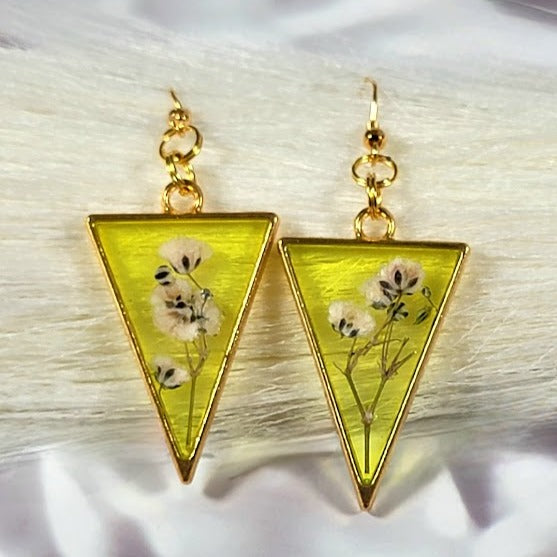 Bright yellow, triangular resin earrings featuring delicate white flowers within, framed by gold trim, present a botanical charm on a plush backdrop.