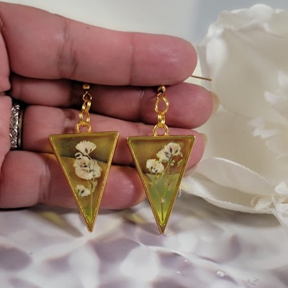 A hand delicately presents yellow resin triangle earrings with preserved white flowers, hanging from gold hooks against a rippling satin background.