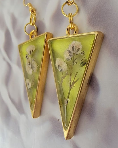 Golden triangular resin earrings with delicate white florals inside, dangling gracefully on golden hooks against a silky backdrop.