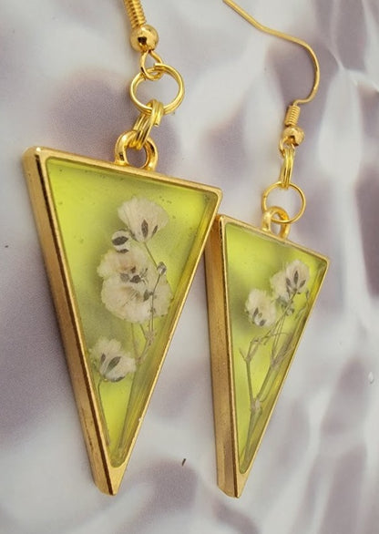 Golden hooks suspend translucent yellow triangular resin earrings, each with a white floral interior, set against a gently draped satin background.