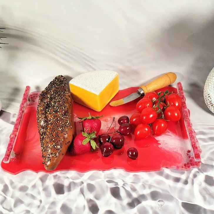 Bright coral-colored resin tray with translucent edges showcasing encased pink particles adorned with fresh fruits, bread, and a knife. It is an art piece perfect for lively gatherings.