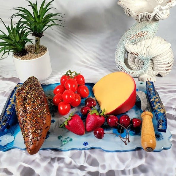 Handcrafted blue resin tray with floral inclusions presenting fresh fruits, a colorful loaf, and a cheese wedge. Paired with a mermaid decor and a potted plant.