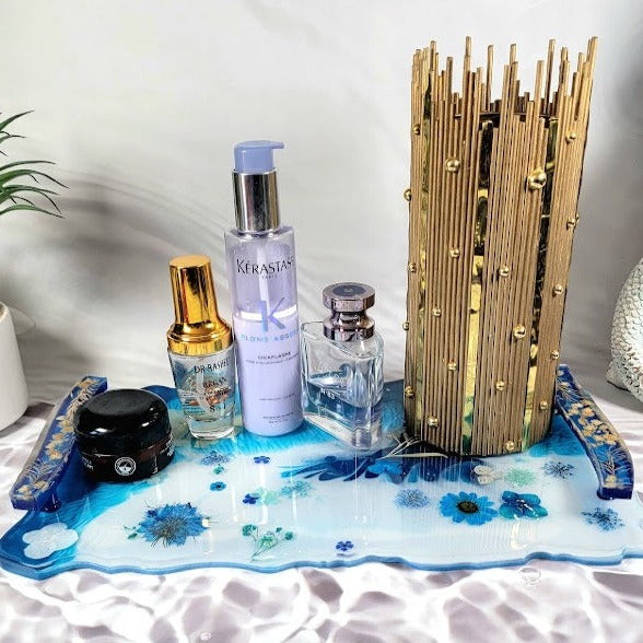 Blue resin tray adorned with real flowers and gold accents displaying an array of beauty products. A seashell sculpture and potted plant enhance the elegant setup.