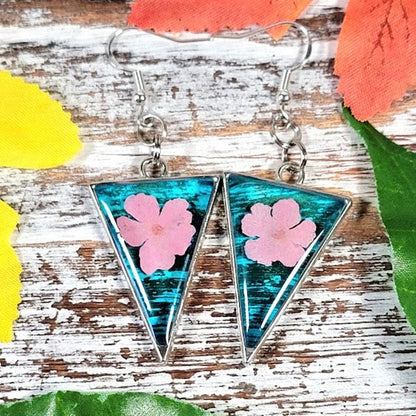 Handcrafted triangular resin earrings suspended from silver hooks, featuring a vibrant turquoise and black abstract pattern with delicate pink floral inlays. 