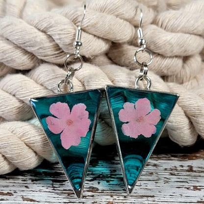 Handcrafted triangular resin earrings showcasing an ombre of turquoise hues with delicate pink floral inclusions, presented on a rustic white and brown wooden background flanked by vibrant autumn leaves.