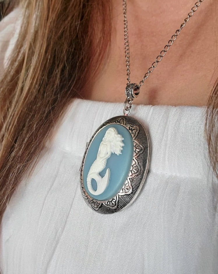 A mermaid cameo pendant necklace hanging on a silver chain that elegantly rests against a woman wearing a white top, its intricate detailing and soft blue hue adding a touch of mythical grace.